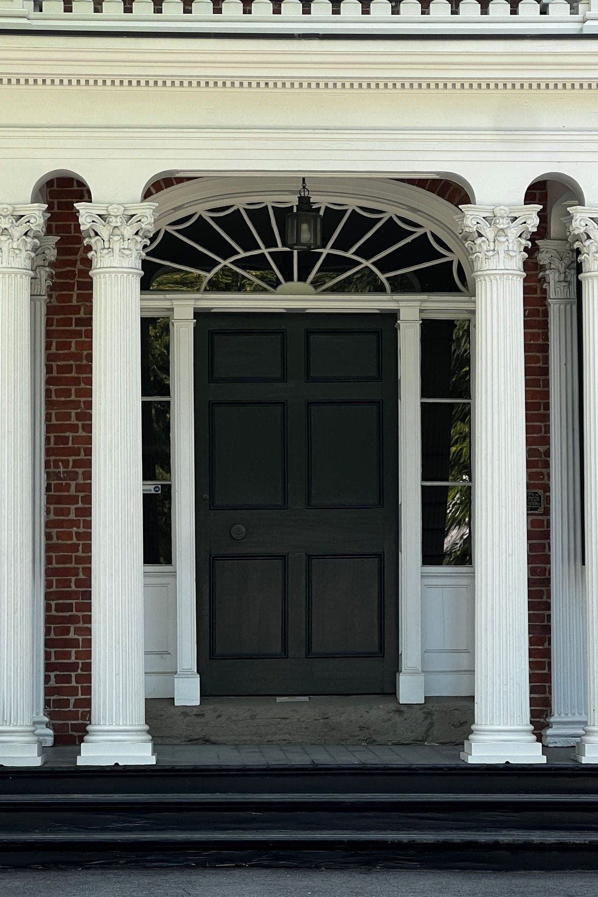 The front door of an older brick house, with white columns, black shutters and a fanlight window above the door.